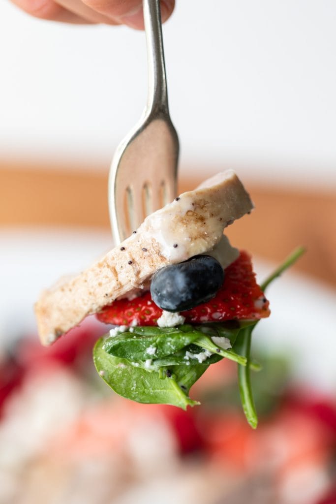 Fork holding piece of chicken as well as blueberry, strawberry, and piece of spinach with salad in background