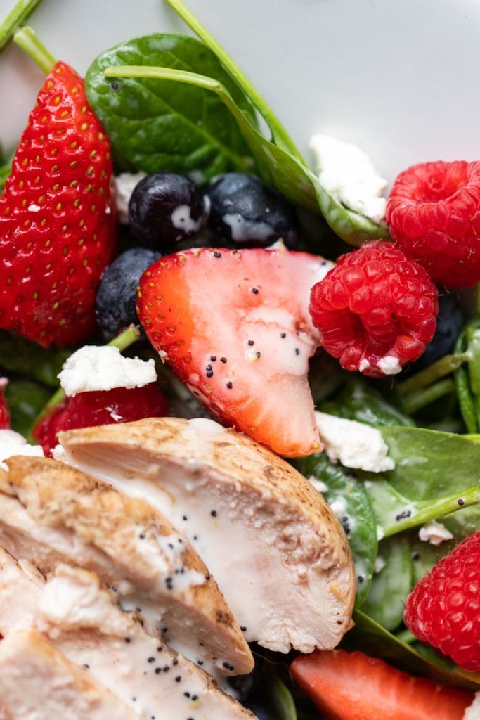 Half piece of strawberry and raspberries and blueberries sitting on spinach with grilled chicken all on white serving platter