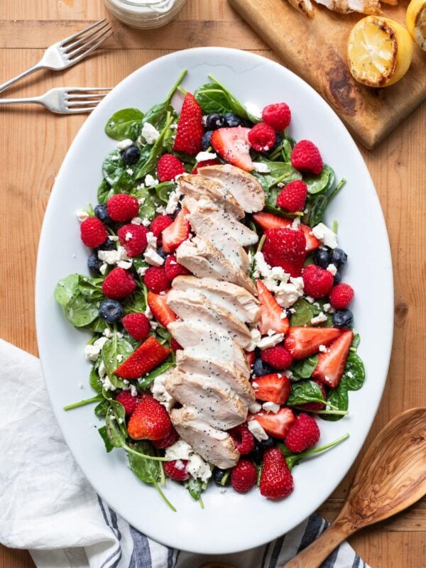 Top down view of grilled chicken berry salad topped with lemon poppy seed dressing served on white platter with extra lemons and dressing all on wood surface