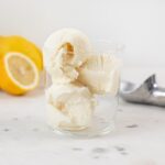 Three scoops of lemon thyme ice cream sitting in glass container with sliced lemon in background and ice cream scoop all on marble surface