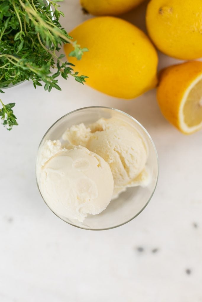 Top down view of lemon thyme ice cream in glass container with lemons and thyme in background all on marble surface