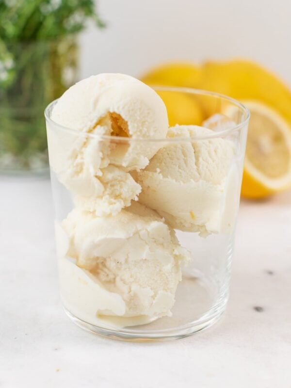 Three scoops of light yellow ice cream sitting in glass on marble surface with lemons and thyme in background