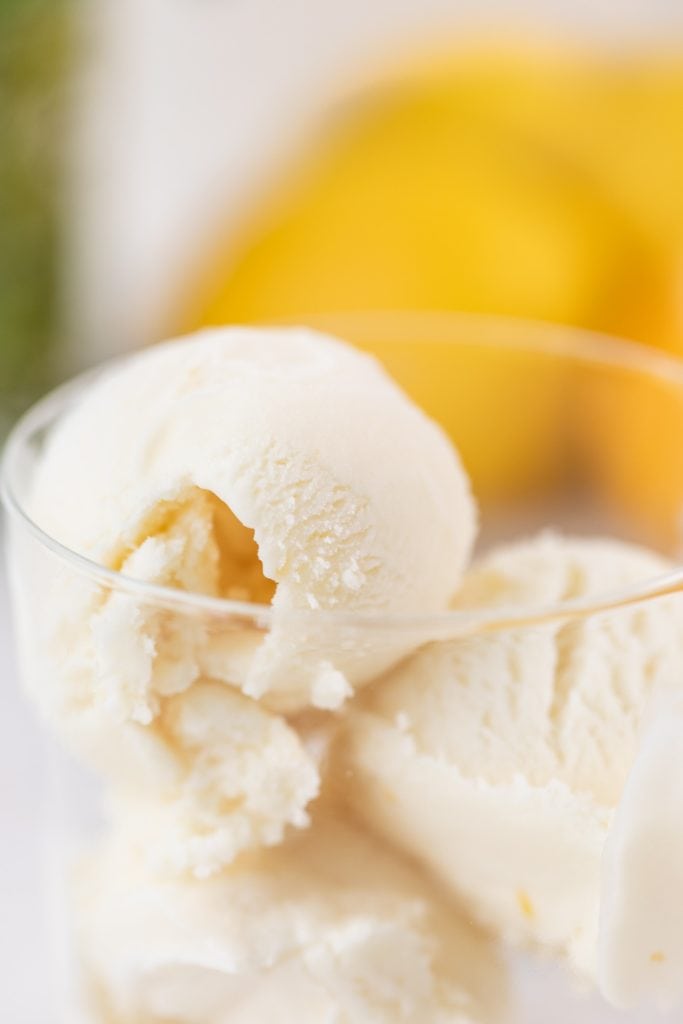 Curled scoop of light yellow ice cream sitting in glass container with blurry lemons in background all on white marble surface