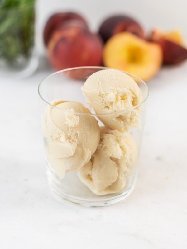 Glass full of three scoop of peach rosemary ice cream with extra peaches and rosemary in background all on marble surface