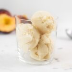 Three scoops of ice cream sitting in a glass with peaches and ice cream scoop in background all on marble surface