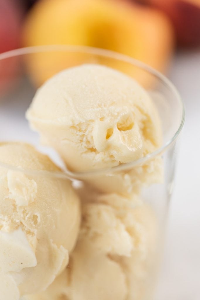 Close up view of light yellow scoops of ice cream sitting in glass with peach slices in background all on marble surface