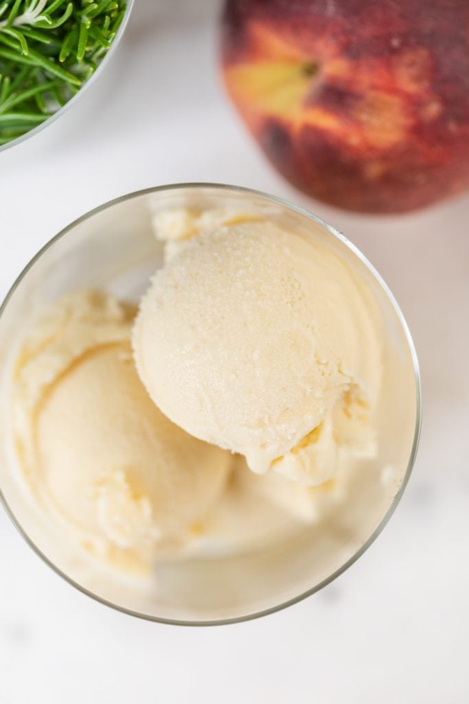 Top down view of scoop of ice cream in a glass container with peaches and herbs sitting on marble surface