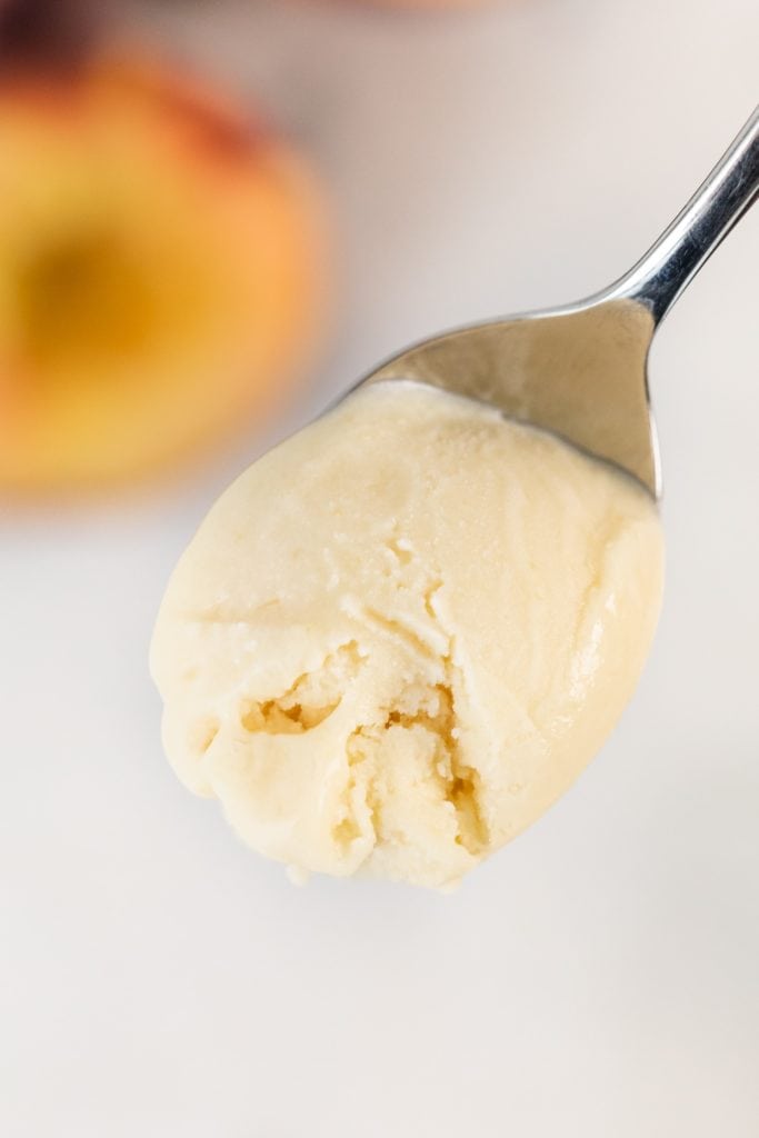Spoon holding peach rosemary ice cream with slice of peach in background sitting on marble surface