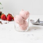 Three scoops of pink-colored strawberry basil ice cream sitting in glass with peppercorns, strawberries, and ice cream scoop on background all on piece of white marble
