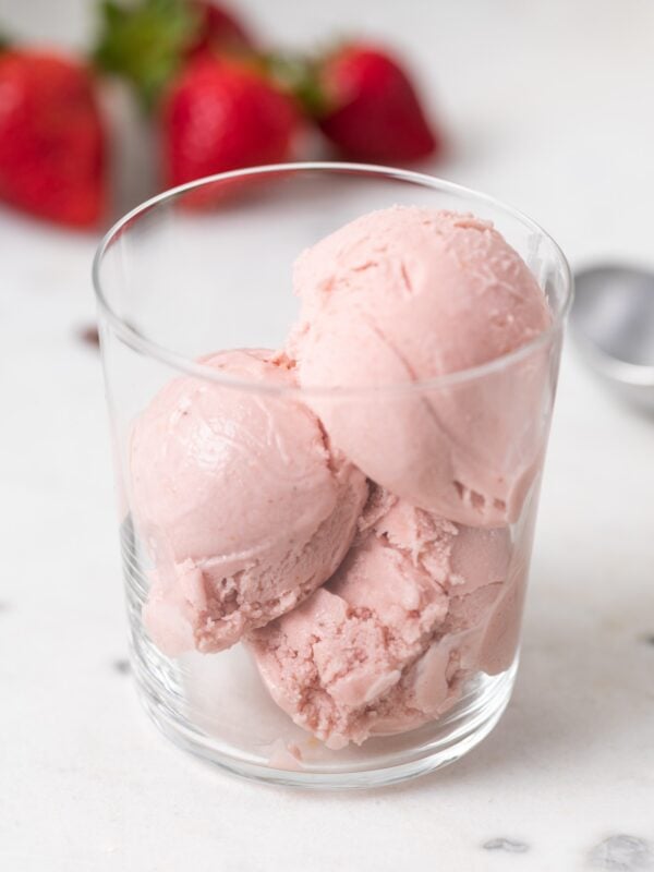 Three scoops of pink-colored strawberry basil ice cream sitting in glass cup with strawberries in background all on marble surface