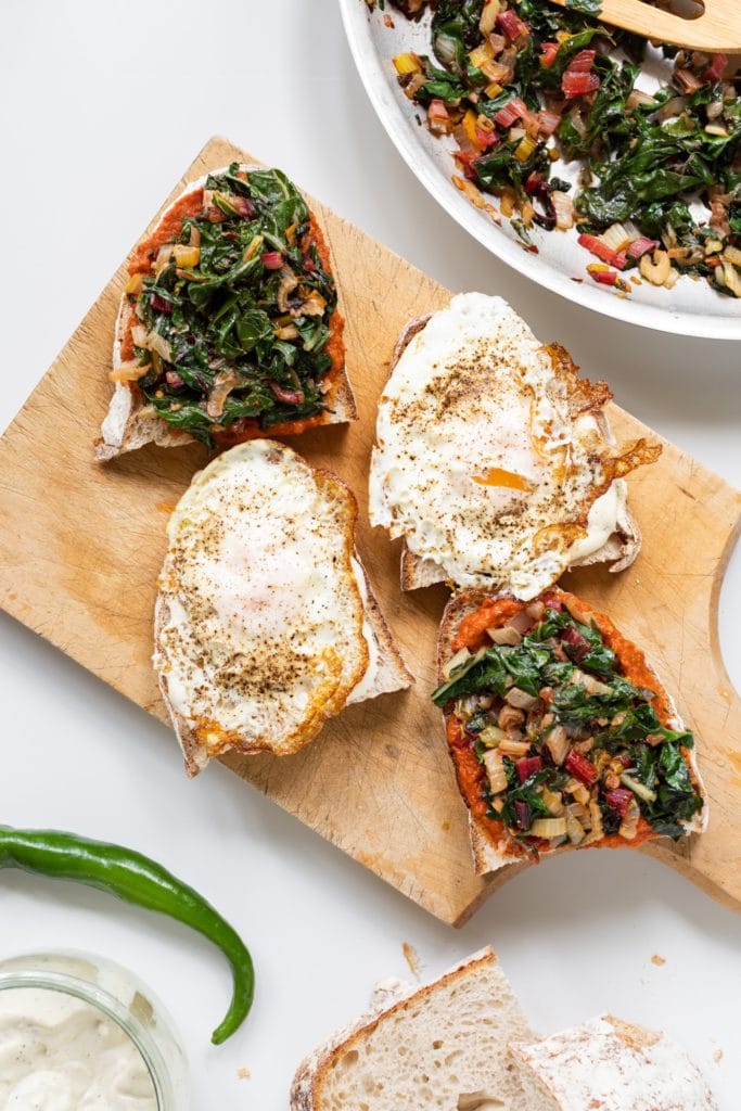 Top down view of open faced sandwiches topped with fried eggs and Swiss chard mixture sitting on wood cutting board with extra pieces of bread all on white surface