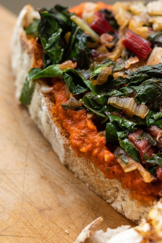 Harrisa sauce on piece of sourdough bread topped with Swiss chard mixture sitting on wood cutting board