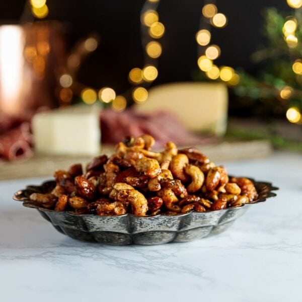 Bowl of candied nuts sitting on white piece of marble with twinkling holiday lights in background