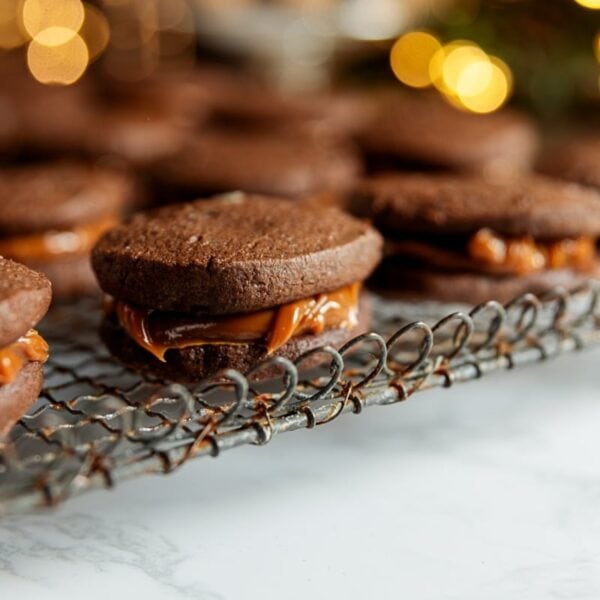 Chocolate sandwich cookie with creamy dulce de leche inside resting with more cookies all on wire cooling rack on white marble surface.