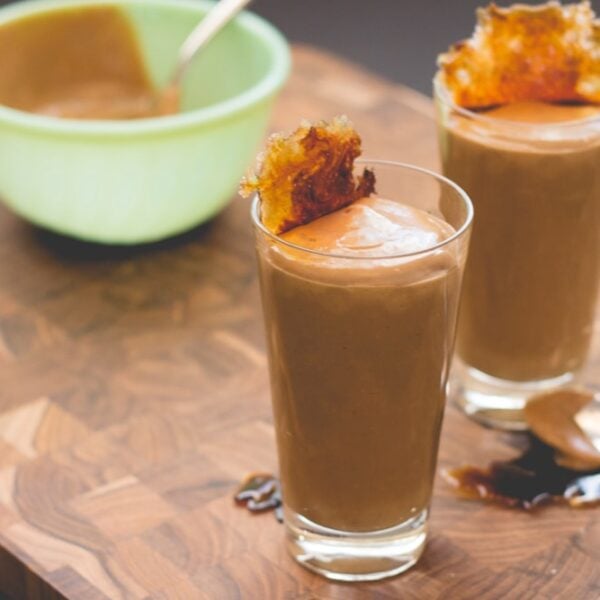 Tall glass filled with brown colored pudding with browned sugar crisp on top with light green bowl sitting in background all on wood cutting board surface