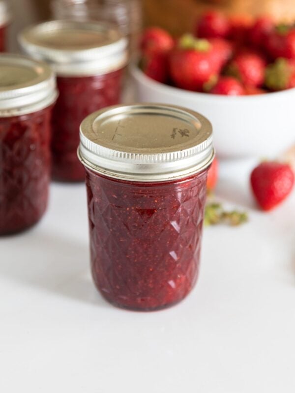 Glass jar filled with red colored strawberry jam with bowl of strawberries sitting in background along with a large copper kettle