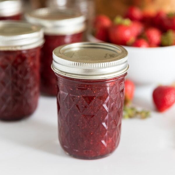 Glass canning jar sitting on white countertop with white bowl filled with strawberries with copper kettle in the background