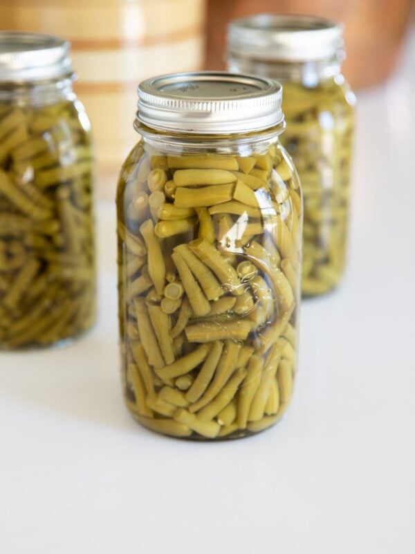 Glass jars filled with sliced green beans with lids and ring on top sitting on white countertop with copper pot in background