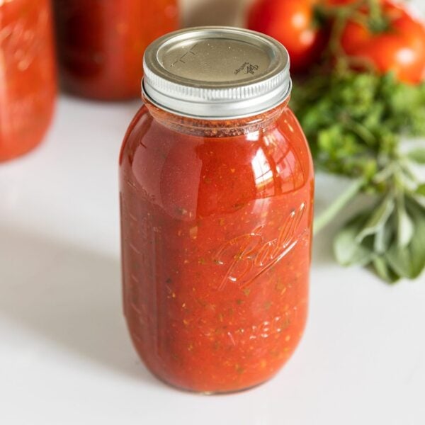 Glass jar filled with red spaghetti sauce sitting on white countertop with green herbs and red tomatoes in background with extra glass jars filled with sauce