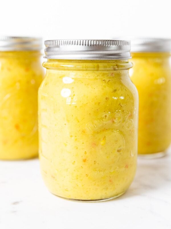Glass canning jars filled with yellow mustard liquid with bits of peppers showing all on white marble surface.