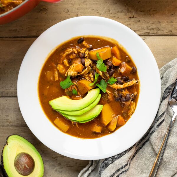Top down view of white bowl filled with red colored chili with slices of avocado on top with extra avocado and soup sitting on wood table