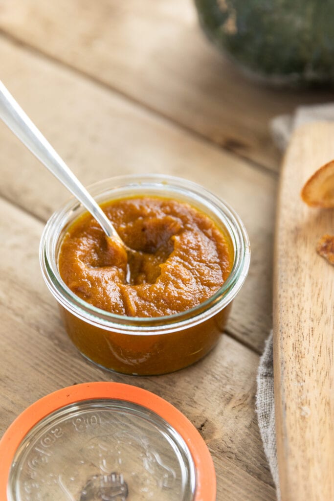 Glass jar holding brown colored spread with spoon in mixture on top of wood table with pumpkin in background