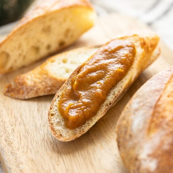 Slice of French bread spread with brown colored pumpkin apple butter with extra bread pieces around sitting on wood cutting board