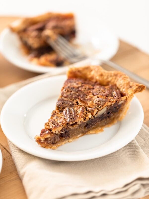 Slice of pecan pie with chocolate layer on bottom sitting on white plate with napkin underneath with half eaten piece of pie in background all on wooden surface