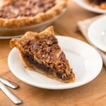 Piece of chocolate bourbon pecan pie with golden crust sitting on white plate with forks beside with extra pieces of pie sitting in background along with rest of pie all on brown wooden surface