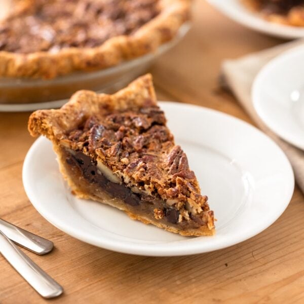 Piece of chocolate bourbon pecan pie with golden crust sitting on white plate with forks beside with extra pieces of pie sitting in background along with rest of pie all on brown wooden surface