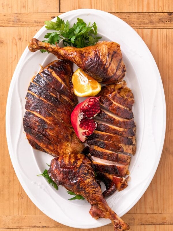 Top down view of citrus dry-brine turkey pieces sitting on white serving platter with pomegranate and orange slice all on wood board