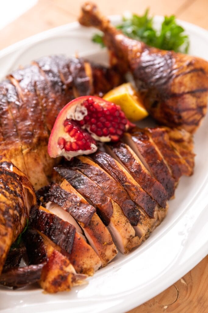 Slices of citrus dry-brine turkey sitting on white platter with turkey legs sitting beside with pomegranate and orange slice all on wood board