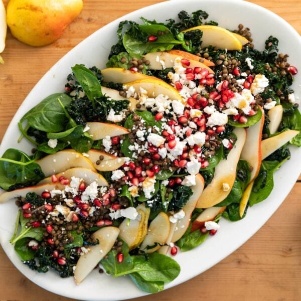 Top down view of white platter sitting on wood board containing a hearty kale salad topped with pears, pomegranate arils, lentils, and crumbles of feta cheese