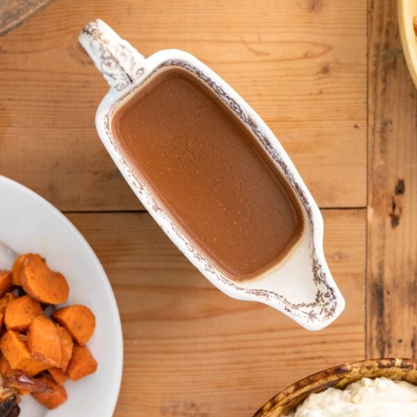 Top down view of brown colored homemade gravy in a white gravy container with extra food sitting on white plate and extra side dishes around all on wood surface