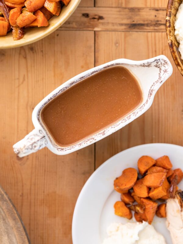 Top down view of gravy boat holding brown colored homemade gravy surrounded by plate filled with Thanksgiving food and extra carrots all on wood surface
