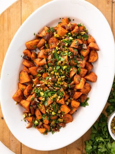Top down view of orange roasted carrots sitting in white serving platter topped with dates and pistachios all on a wood surface with extra parsley