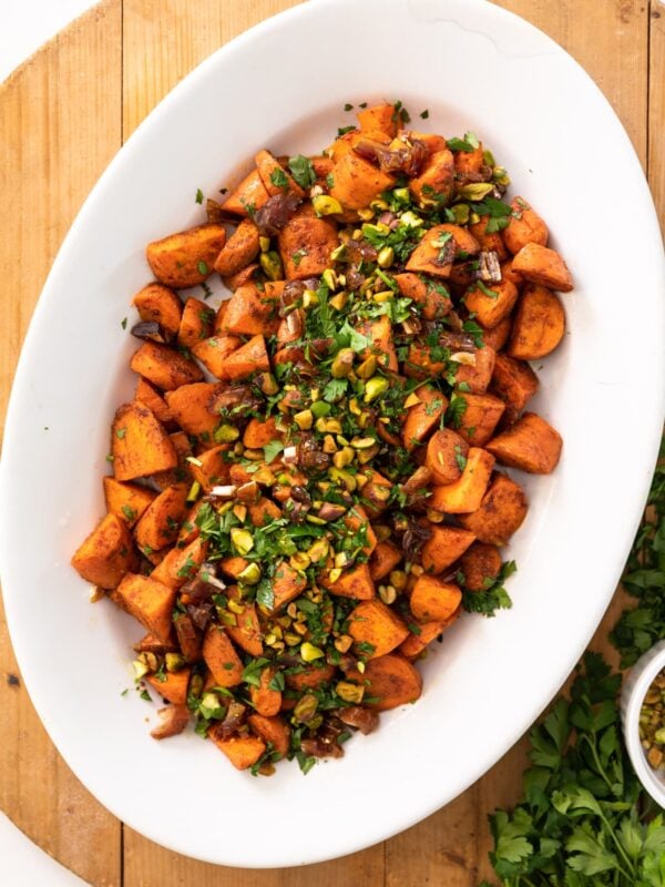 Top down view of orange roasted carrots sitting in white serving platter topped with dates and pistachios all on a wood surface with extra parsley