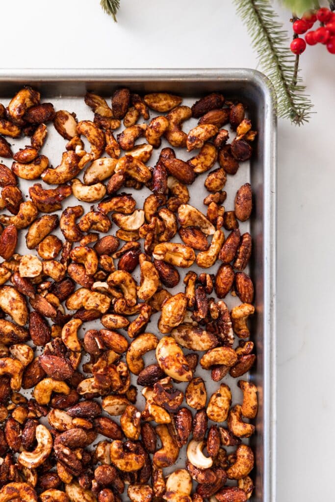 Top down view of cinnamon spiced nuts sitting on silver baking pan on white countertop with Christmas greenery and red berries to the side
