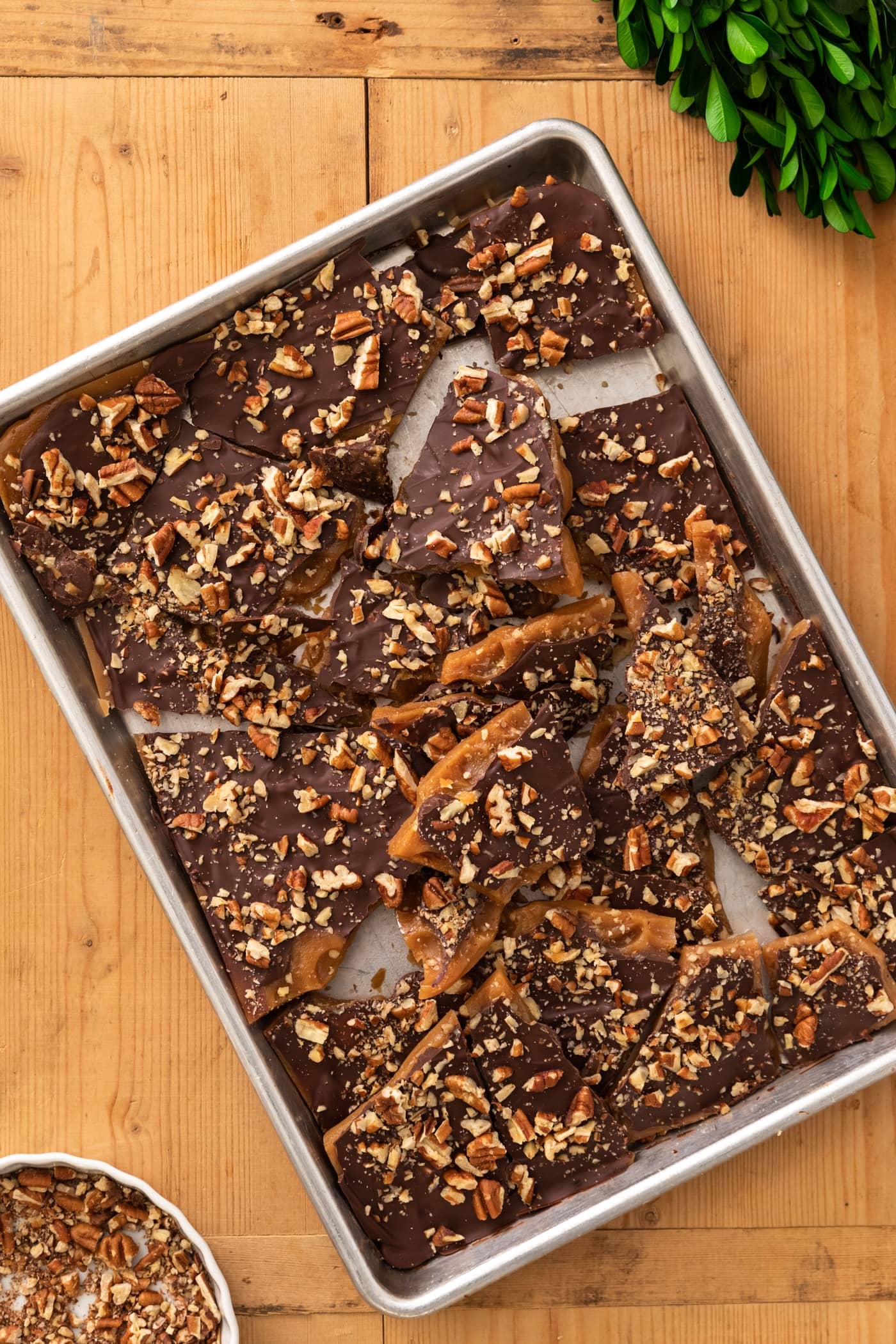 Top down view of silver baking pan filled with broken pieces of toffee covered with chocolate and nuts with pan of chopped nuts sitting on wood surface
