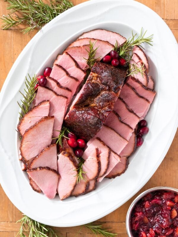 Top down view of pink pieces of ham sliced and placed on white serving platter with sprigs of rosemary around along with silver container filled with red chutney