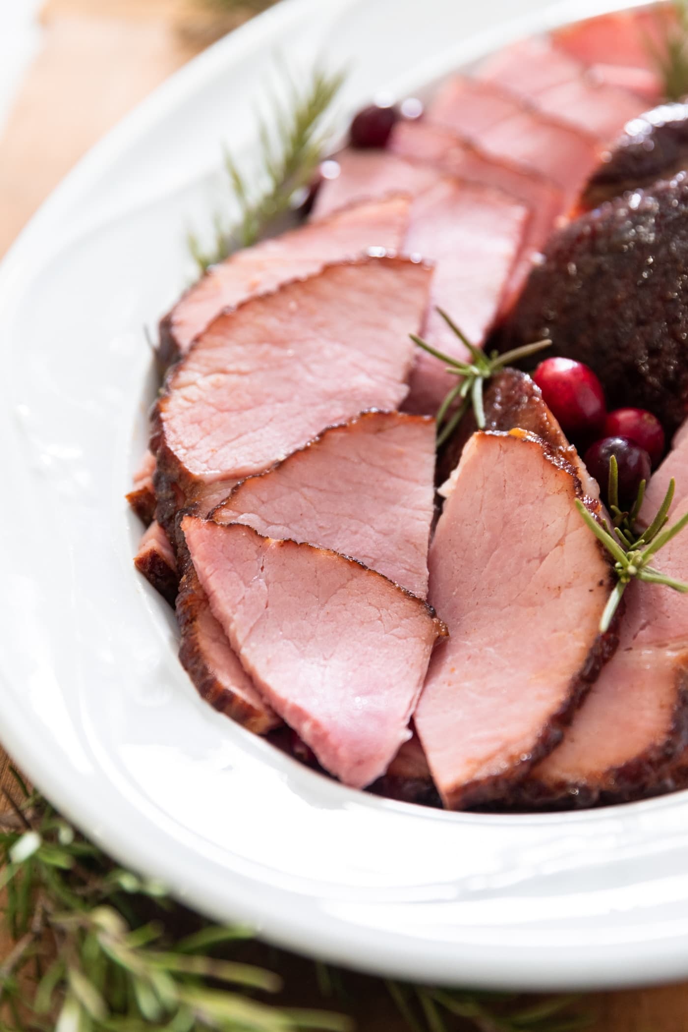 Slices of ham sitting on white platter surrounded by cranberries and sprigs of rosemary all on wood surface