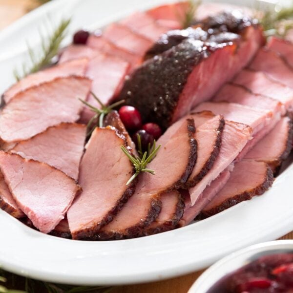 White platter filled with slices of ham along with cranberries and sprigs of rosemary all on wooden board