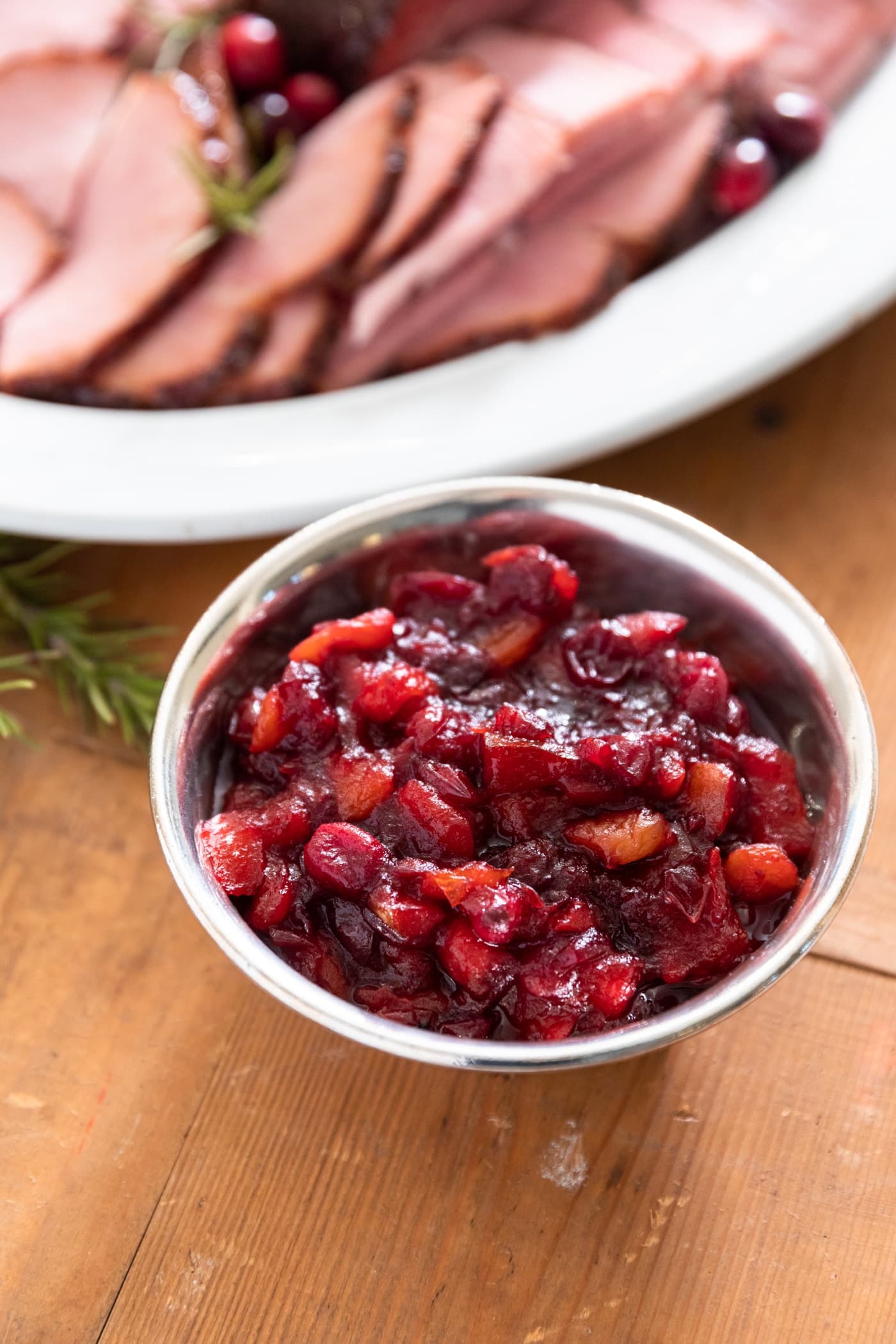 Silver dish filled with red chutney sitting on wood surface with platter filled with slices of ham behind