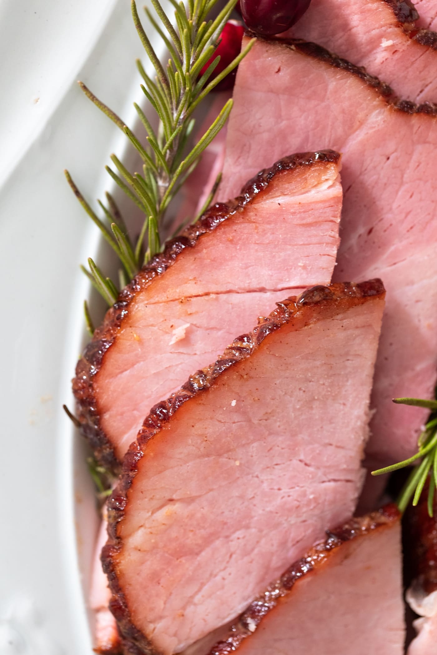 Slices of pink ham sitting on white platter with crispy glaze around edges amidst pieces of rosemary and cranberries