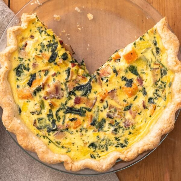 Top down view of yellow quiche sitting in clear glass pie plate dotted with pieces of sweet potato and spinach all on wood surface
