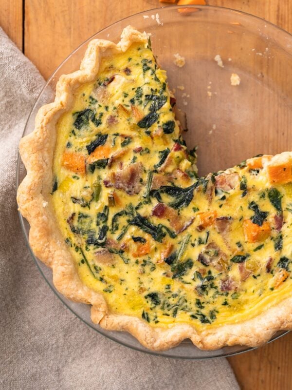 Top down view of yellow colored sweet potato quiche dotted with spinach and pieces of bacon all on wood surface