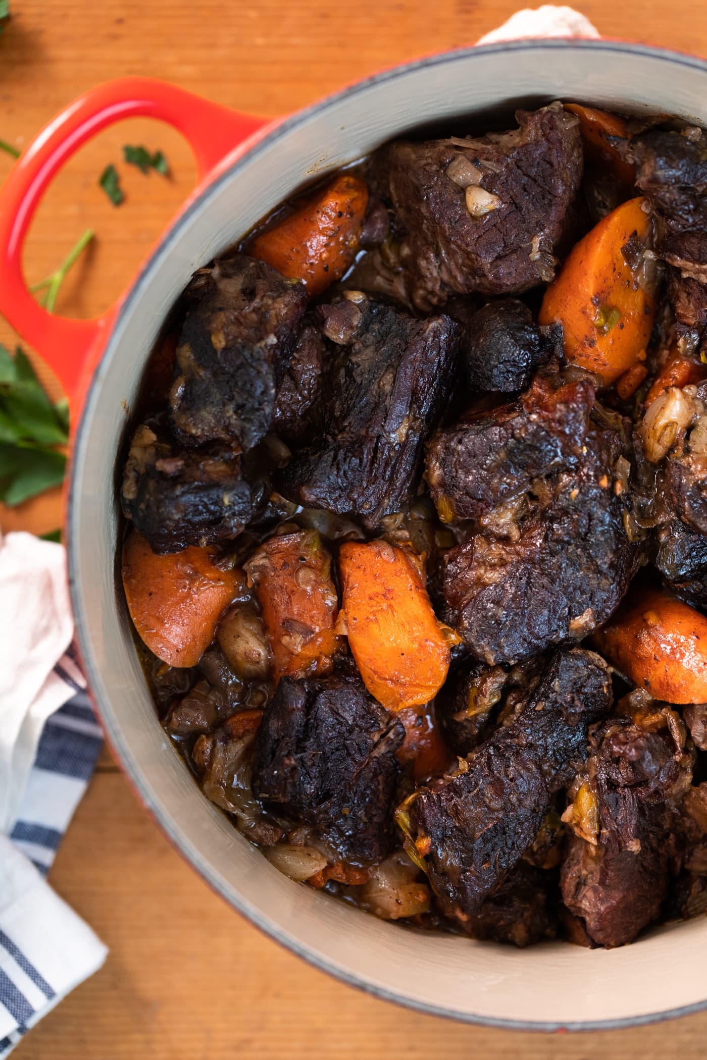 Top down view of red pot filled with pieces of beef and carrots all on wood surface with white napkin