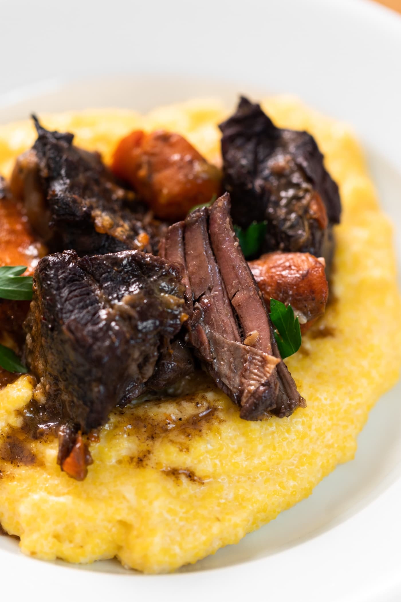 White plate filled with yellow colored polenta topped with pieces of beef and carrots along with parsley