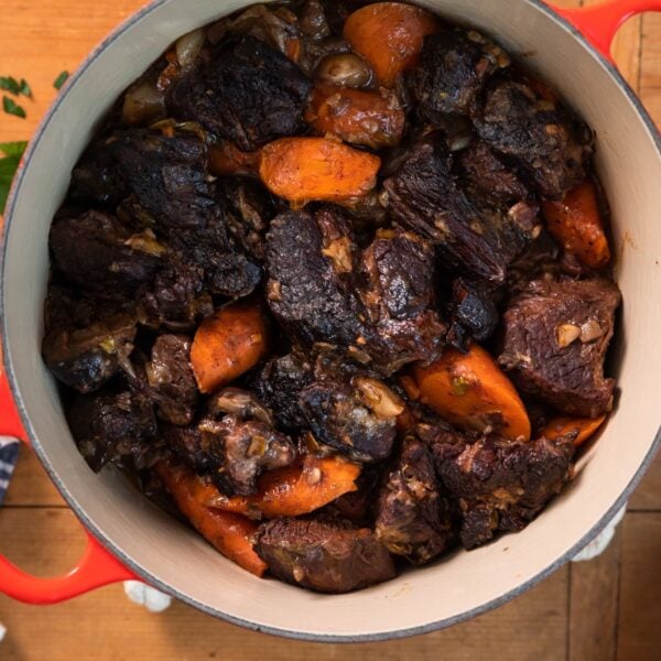 Top down view of red kettle holding pieces of beef and carrots all on wood surface with white plates sitting around