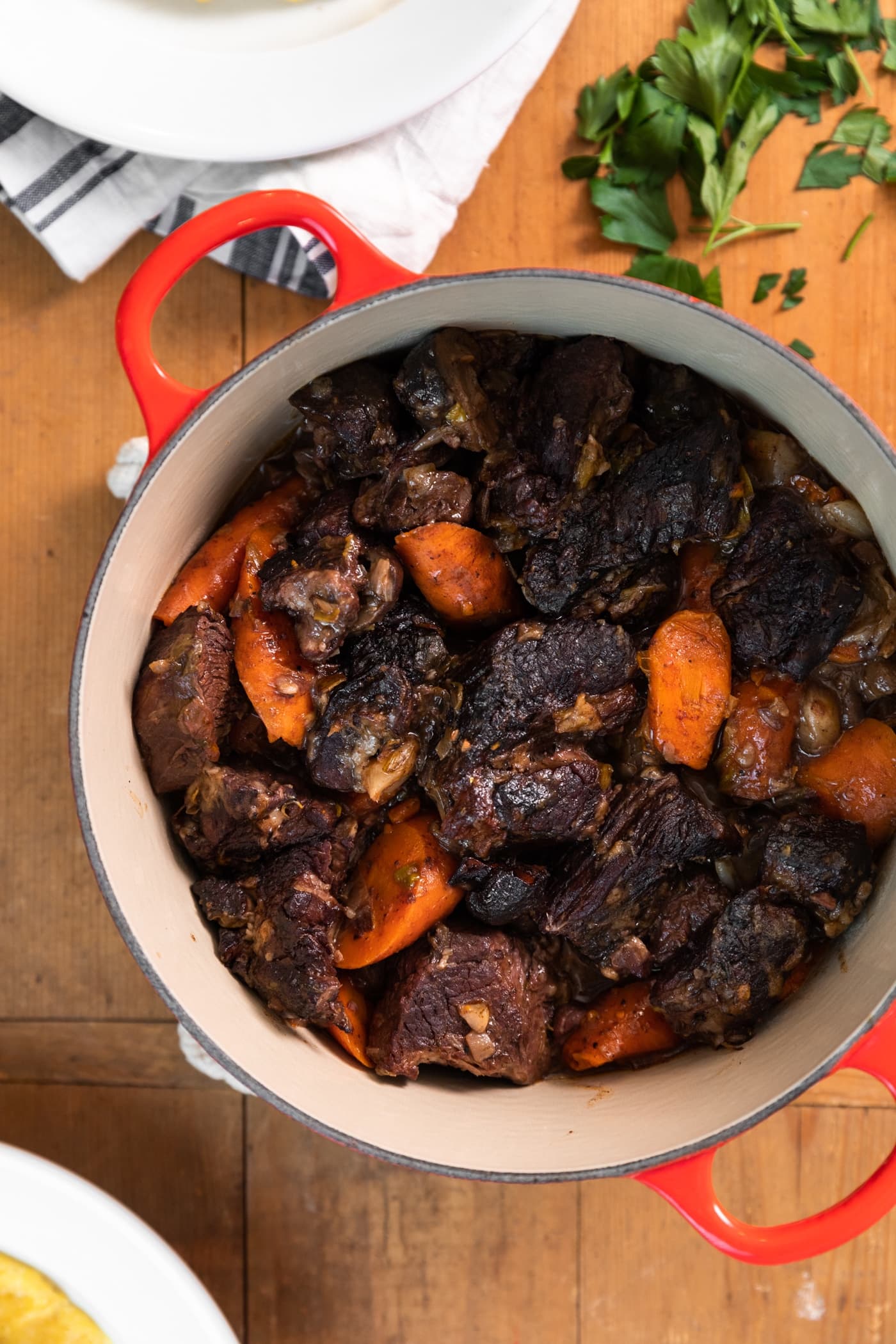 Top down view of red pot filled with pieces of beef with carrots all on wood surface with white plates
